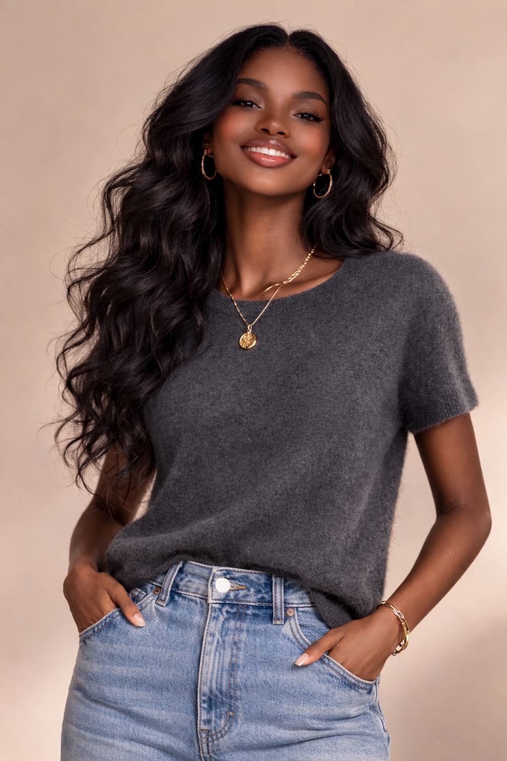 Smiling woman in a charcoal short-sleeve knit top with high-waisted light blue denim, gold hoop earrings and layered necklace, studio background.