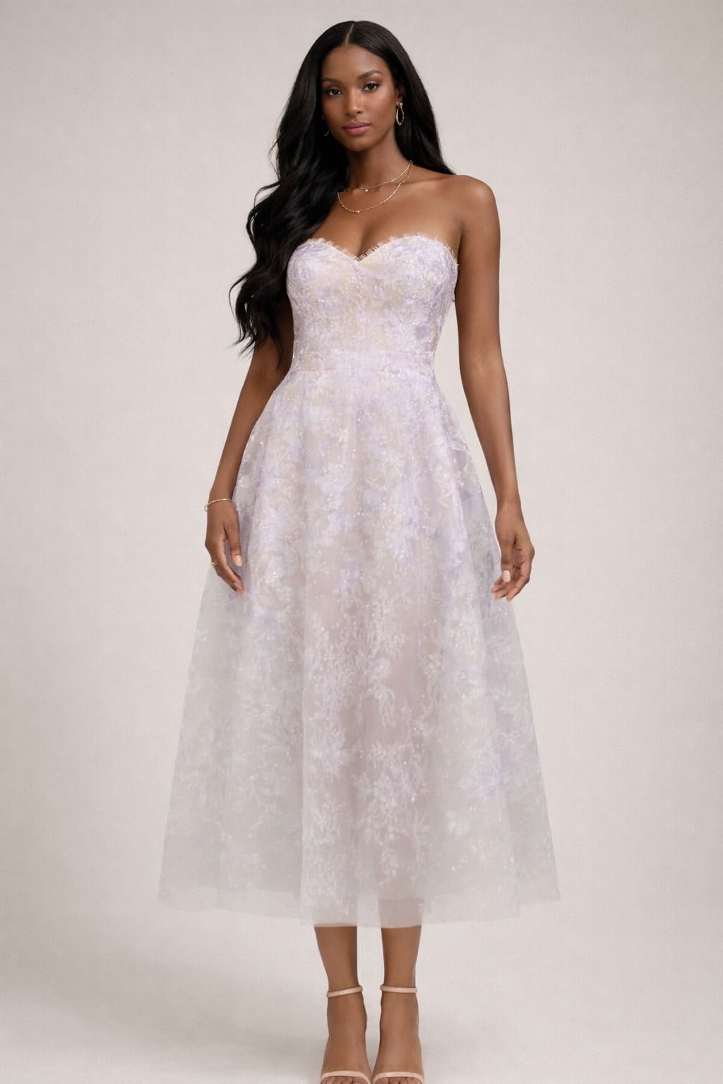 Full-body studio portrait of a Black woman with long wavy hair wearing a strapless lavender tulle midi dress with subtle floral texture and nude strappy heels on a beige background.